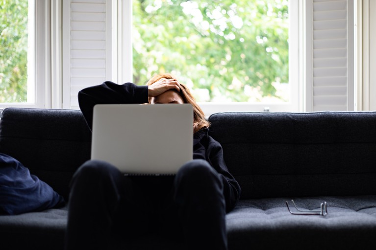 A woman sits on a dark couch with her face obscured by a laptop
