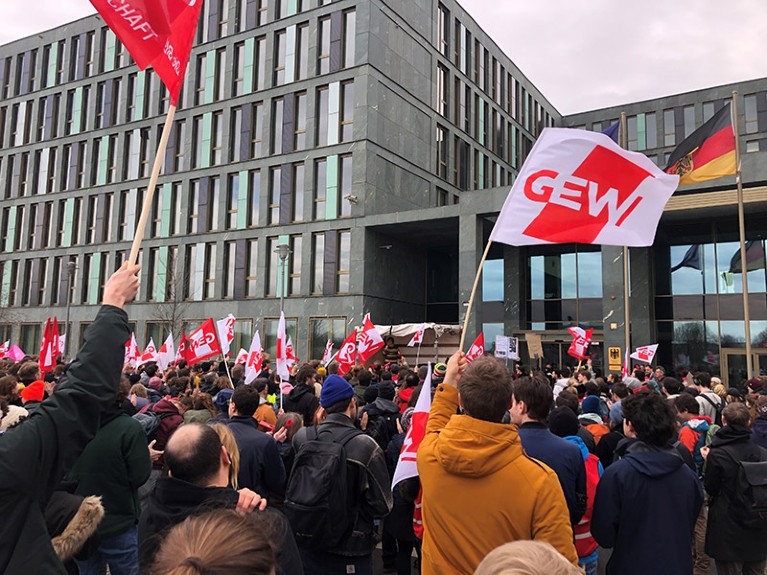 People gather in front of a municipal building waving red and white flags reading 'GEW'.