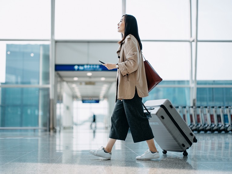 Young Asian woman carrying suitcase and holding smartphone on hand, walking in airport terminal.