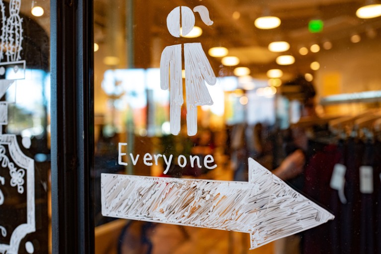 Close-up of hand drawn sign on a window depicting a stick figure which is half female and half male with an arrow