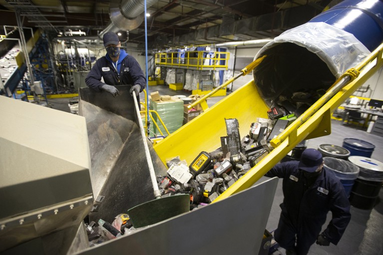 在回收设施中，一名戴着防护面具的工人使用工具将电池推到传送带上