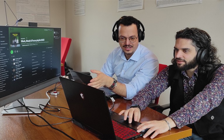 Andrea Caputo and a colleague wear headphones and sit in front of a laptop
