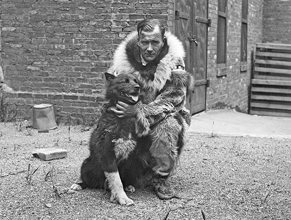 A serious-looking man in Alaska Native fur outerwear is on the ground, his hands around the neck of a small black dog.