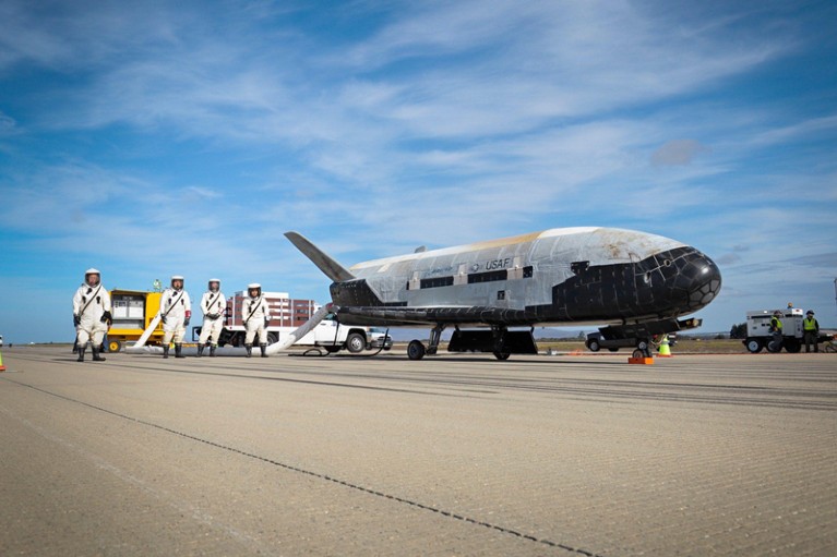 The Boeing X-37B Orbital Test Vehicle and four crew members in white full body suits