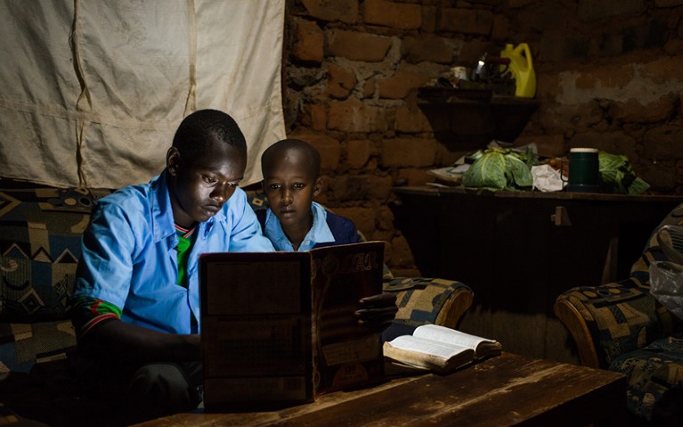 Un escolar, a la izquierda, estudia en casa usando un libro iluminado por un LED, alimentado por tecnología solar M-Kopa.