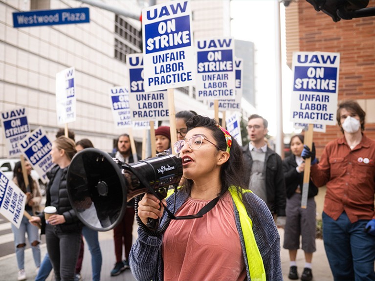 A student holds a megaphone to her mouth and protesters behind her hold picket signs.