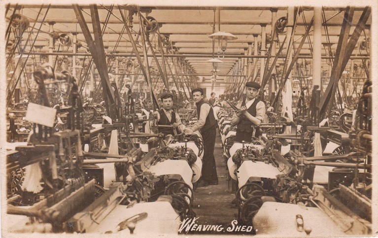 Historic sepia photo of a weaving shed in a cotton mill with three boys and a woman posing between the machinery