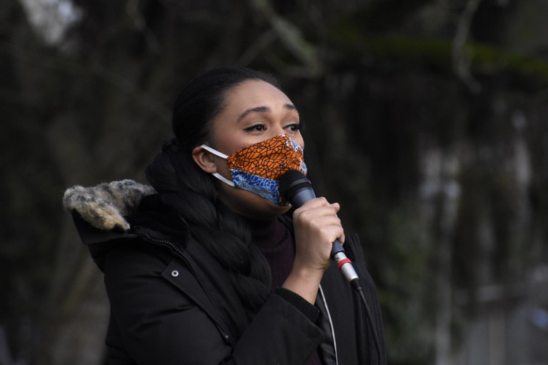 Close-up of Ashley Paynter wearing a face mask and speaking into a microphone with tears in her eyes
