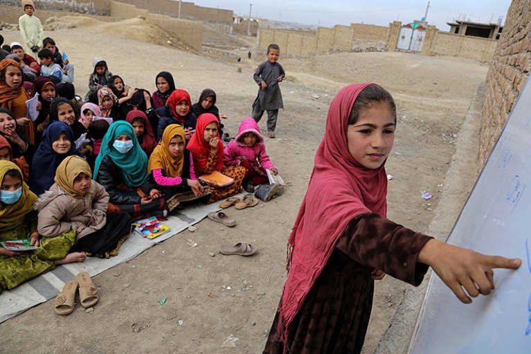 Afghan girl pointing at a whiteboard in an open-air classroom with other children sitting on ground behind her.