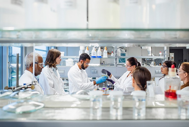 A group of seven scientists of mixed race and mixed gender wearing lab coats and goggles in lab