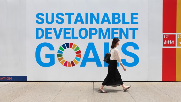 A woman wearing a mask walks past a large sign advertising the UN's Sustainable Development Goals