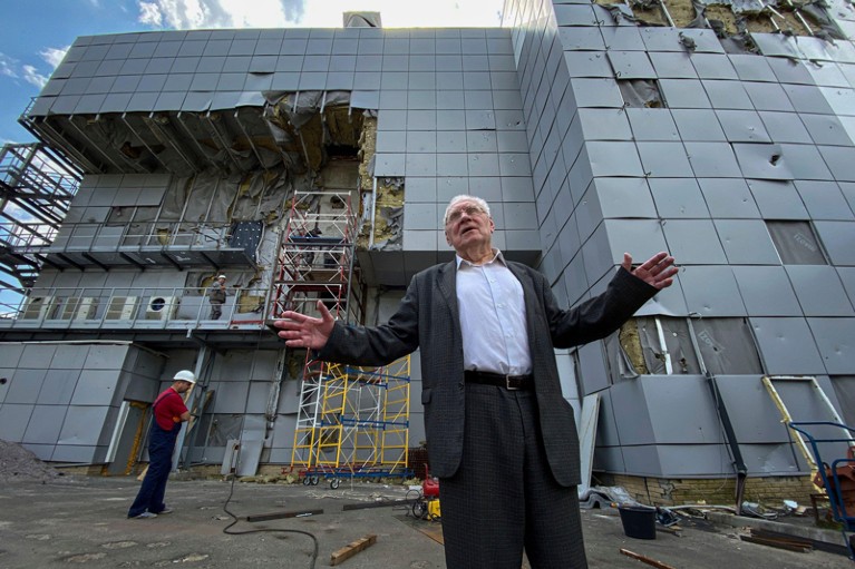 Low-angle view of Mykola Shulga gesturing in front of a damaged nuclear laboratory building