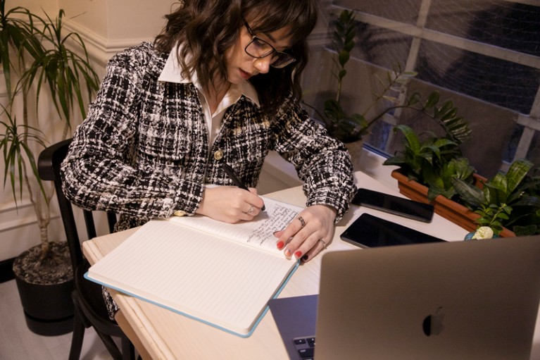 Viktoriia Lemeshchenko-Lagoda sits at a desk in a cafe writing in her notebook with her laptop open in front of her