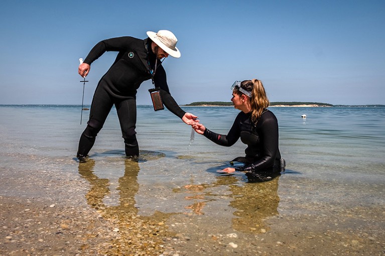 a woman kneeling in shallow water passing a piece of equipment to a man stood next to her