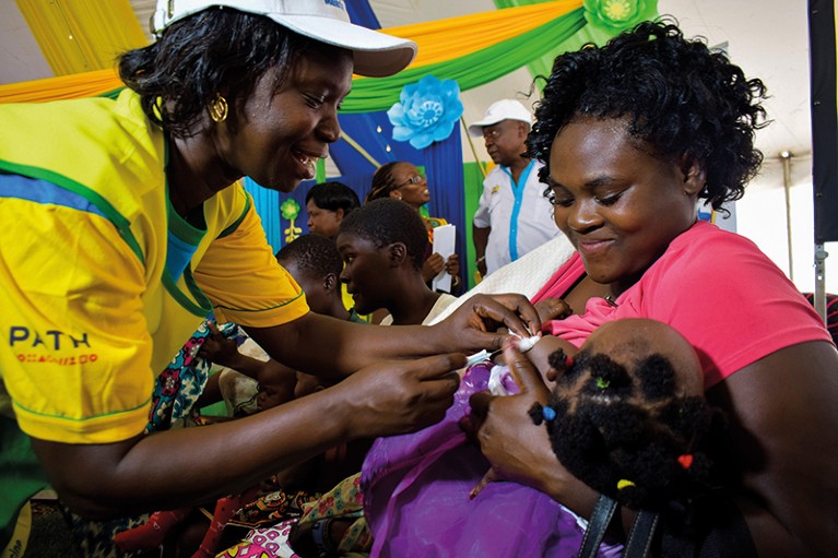 A health worker vaccinates a child in the arms of her smiling mother in a brightly decorated and busy clinic