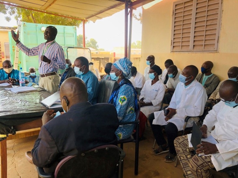 Kayentao gestures as he speaks to unseen community leaders, surrounded by seated medical staff and stakeholders