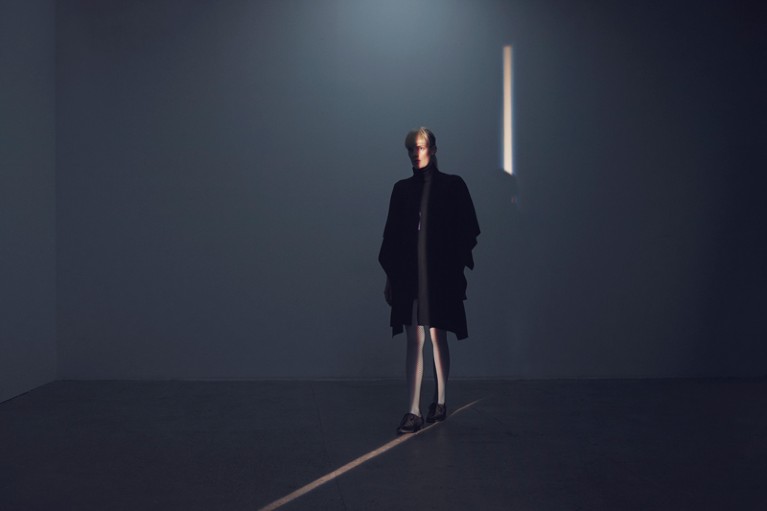 A woman walks along a thin strip of light in a dark concrete room
