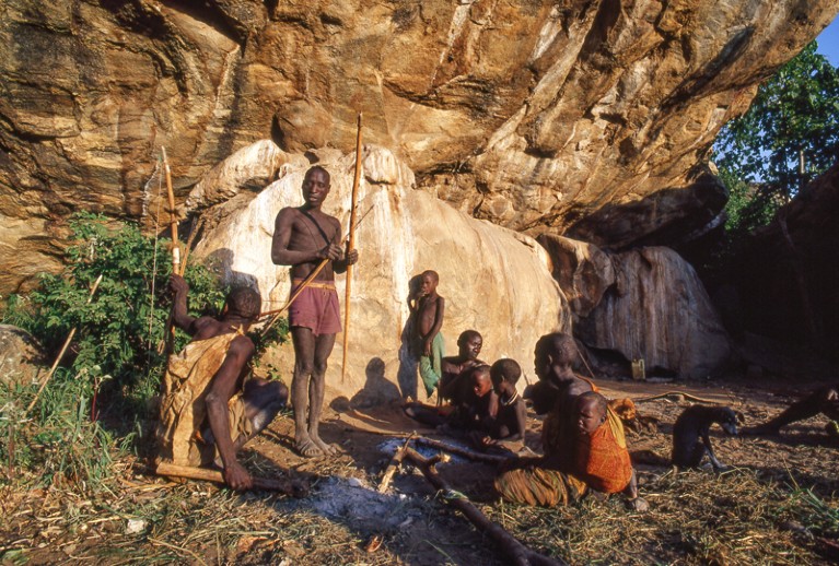 Members of the Hadza people hold large bows and arrows next to a rock face.