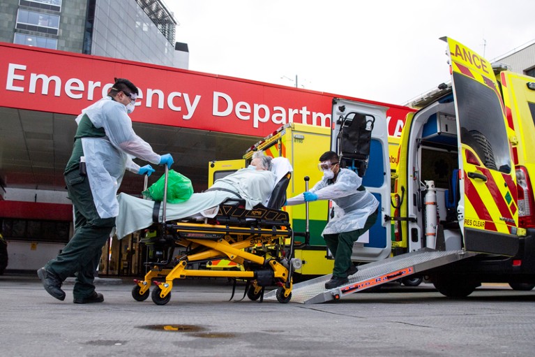 An unwell woman on a stretcher is helped from an ambulance by paramedics wearing PPE at a hospital