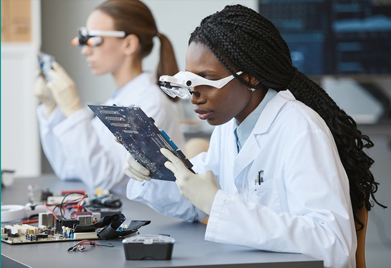Portrait of young black woman wearing magnifying glasses and inspecting electronic parts in quality control lab.