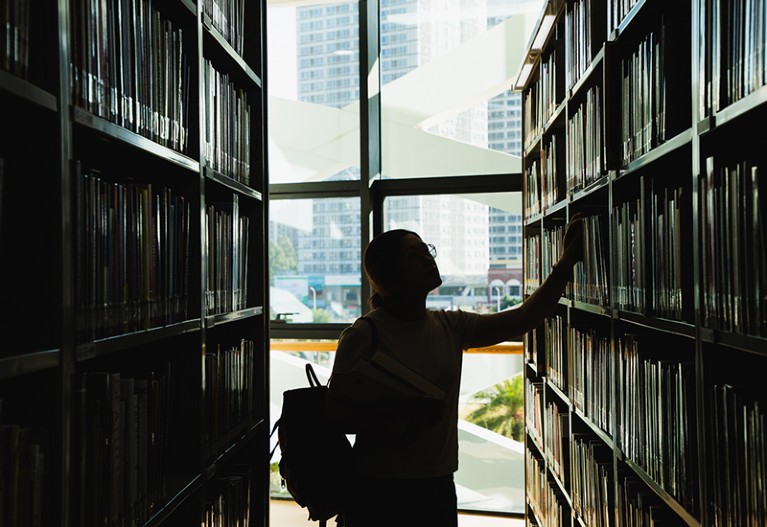 Female student in silhouette looking at the books from the bookshelf.