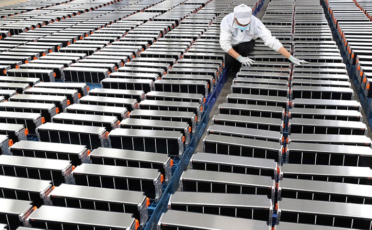 Rows and rows of rectangular silver batteries line a factory floor. A worker wearing white gloves checks the batteries.