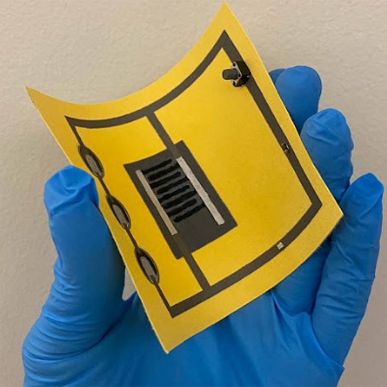 A researcher’s hand wearing blue rubber gloves holds up a paper-thin energy harvesting device