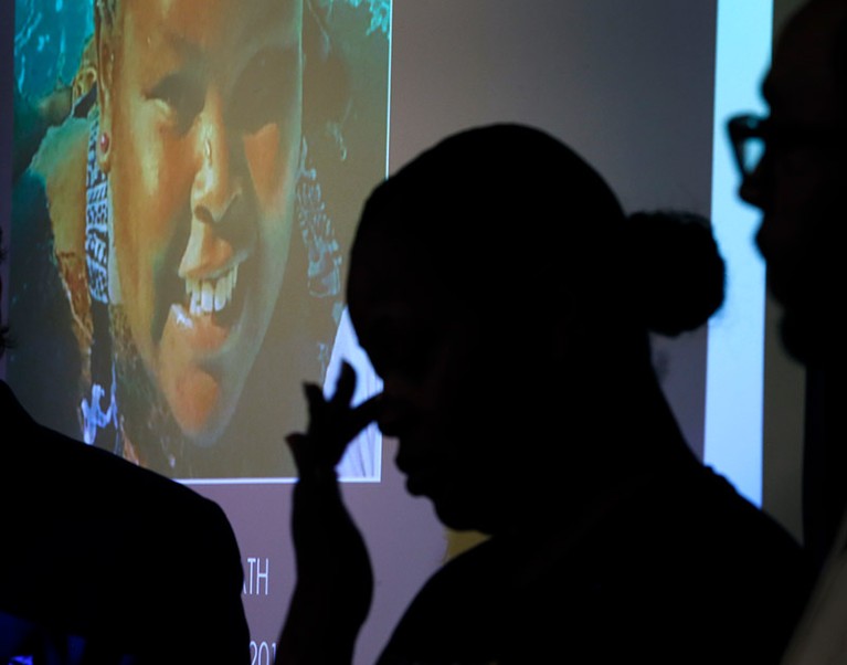A silhouetted Nailah Winkfield wiping tears from her eyes. Behind her is a projected image of her smiling daughter