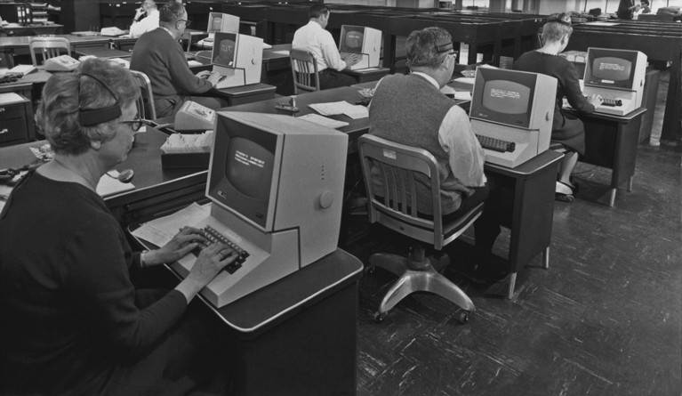 A black and white image of a group of office workers using telephone headsets and computers circa 1965