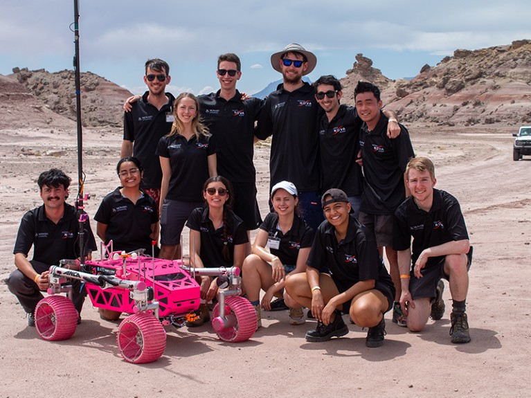 Group of students dressed in black in a desert landscape, with a bright pink rover that they designed at the front.