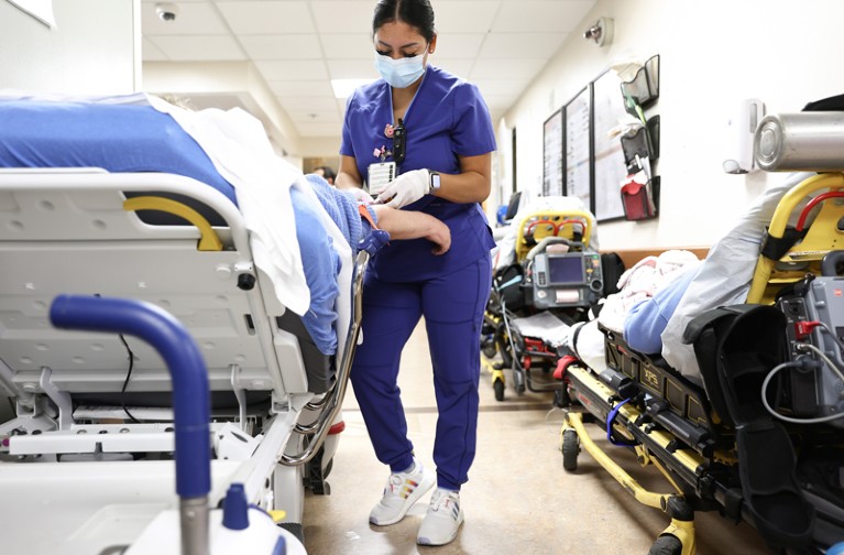 A lab technician wearing a face mask cares for a patient in a hospital bed in the Emergency Department