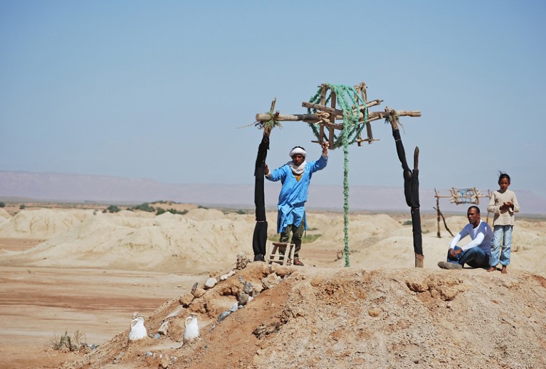 A man extracts water from a khettara in the desert dunes, while a man and child watch