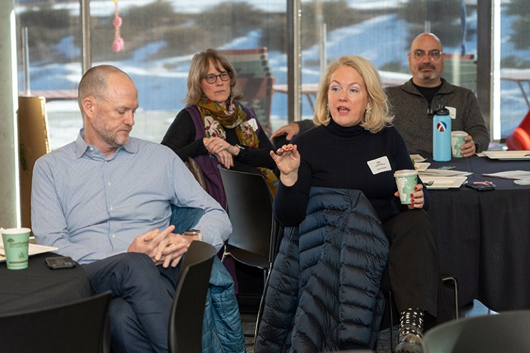 A woman holding a cup speaks among a group of three other people.