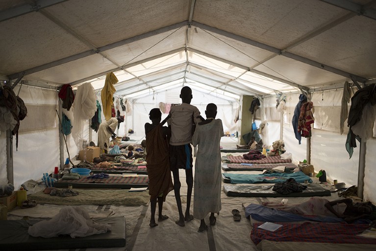 Rear view of a thin man walking the length of a hospital tent supported by two women at either side.