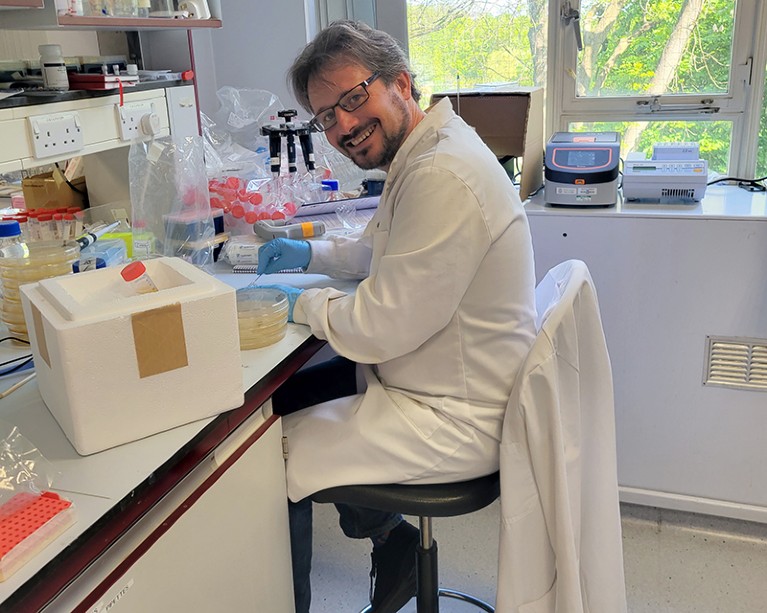 Ian Sudbery sitting at a lab bench wearing a white lab coat.