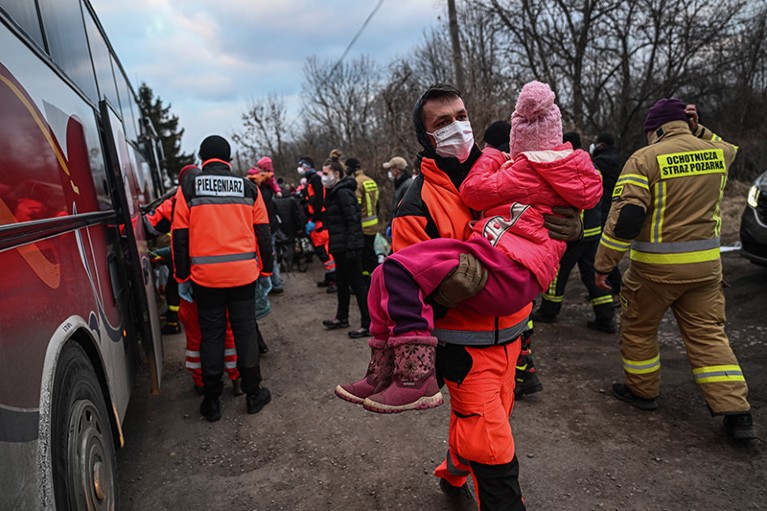 A paramedic wearing a face mask carries a child, a vehicle parked to the left and people stand in the background