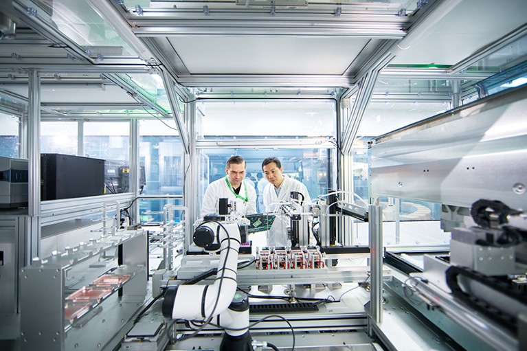 Two people wearing white lab coats stand centrally in a robotics lab, looking at equipment