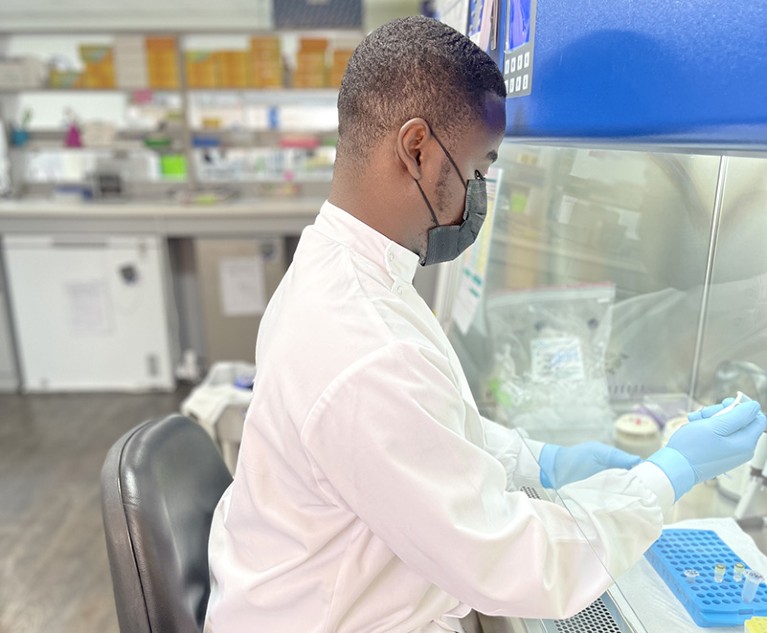 A man wearing white lab coat, gloves, and face mask, sits in a laboratory processing samples