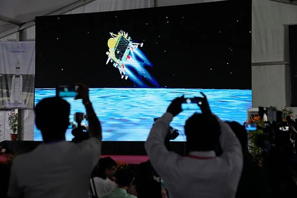Several people use their phones to film an animation of a landing spacecraft displayed on a large screen