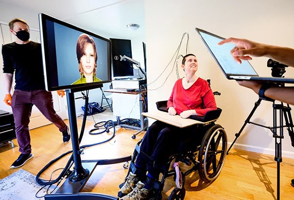 Woman in red shirt connected to machinery sits in wheelchair in front of large screen displaying head of animated avatar.