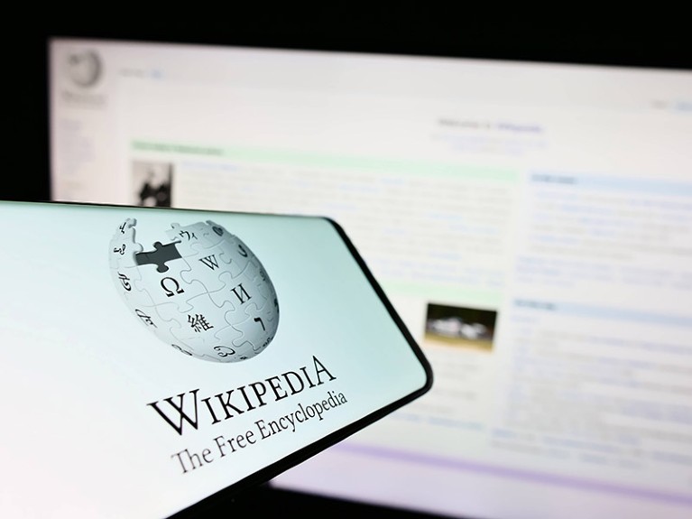 Smartphone with Wikipedia logo on screen held in front of a screen displaying the website.