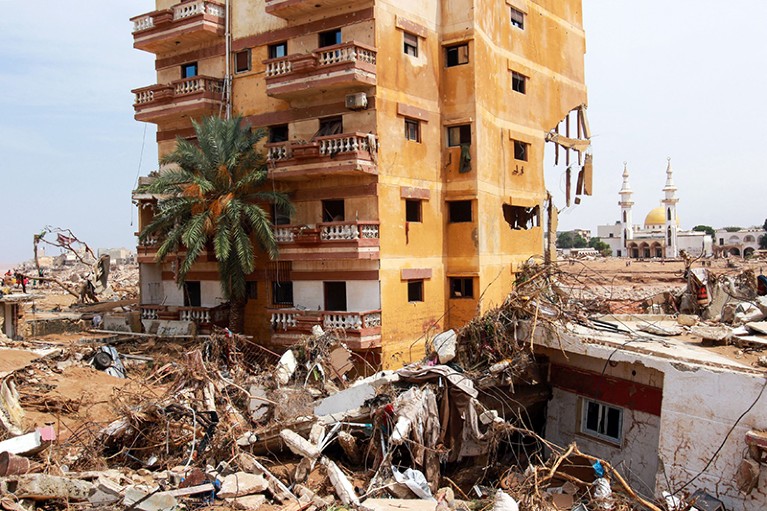A damaged residential block stands among piles of rubble, with an intact mosque in the distance.
