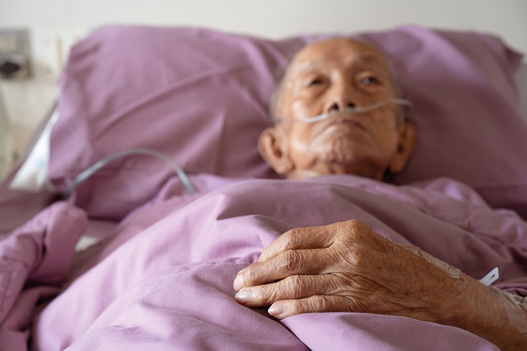 Elderly man lies in bed with purple bedding. He has a nasal oxygen tube. Focus is on his hand resting on the bed.