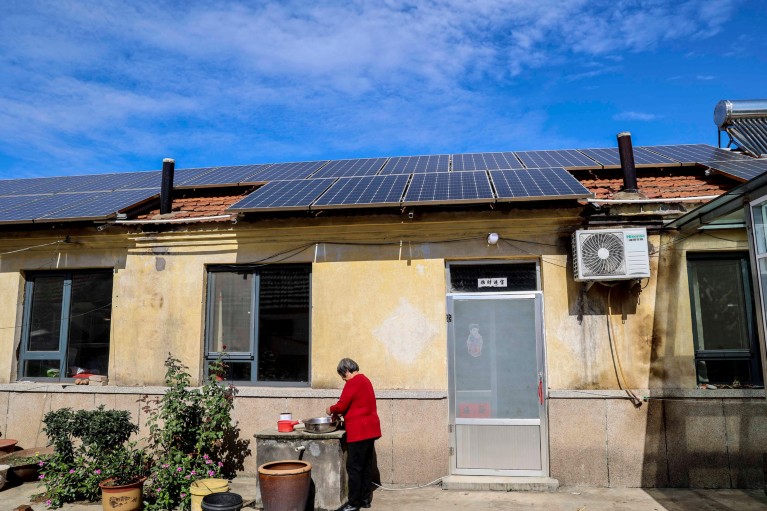 A woman stands with a bowl outside a house with solar panels on the roof.