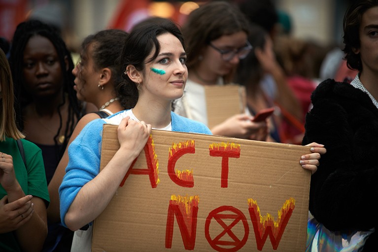 A young woman holds a placard reading 'Act now' with the symbol of Extinction Rebellion.