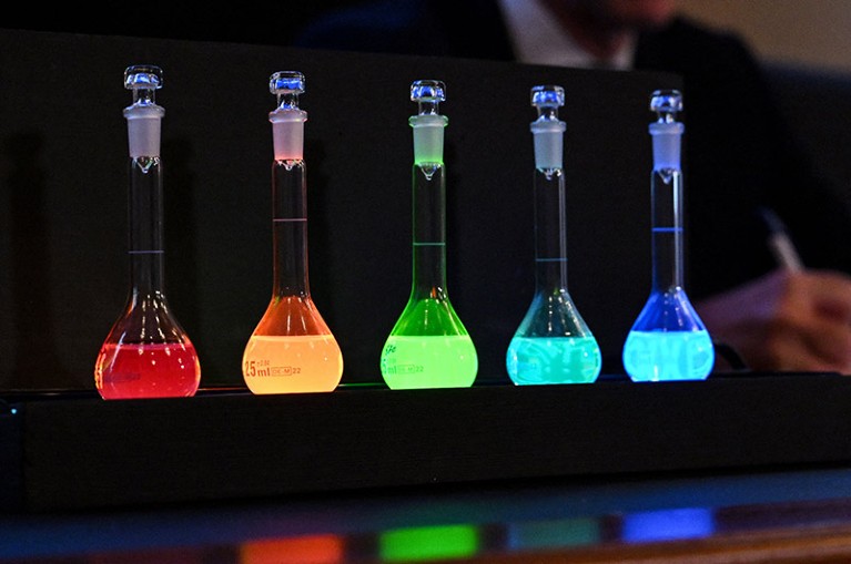 Laboratory flasks in a darkened room, the contents glowing red, orange, green and two shades of blue.