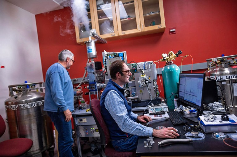 Josh Coon sat at a computer while a colleague in the background tends to nitrogen cooled machinery in the lab