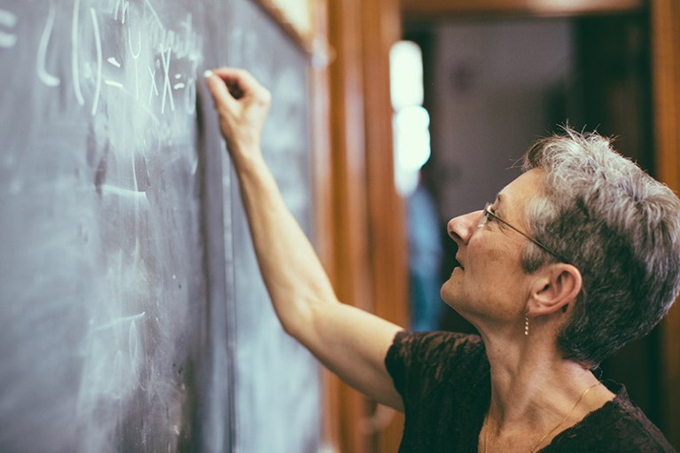 A woman with short, greying hair writing a formula on a chalkboard.