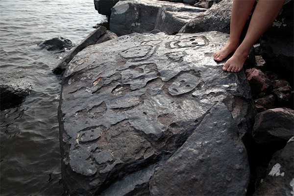 Several rudimentary faces are carved into a large black slab of rock near the water’s edge.