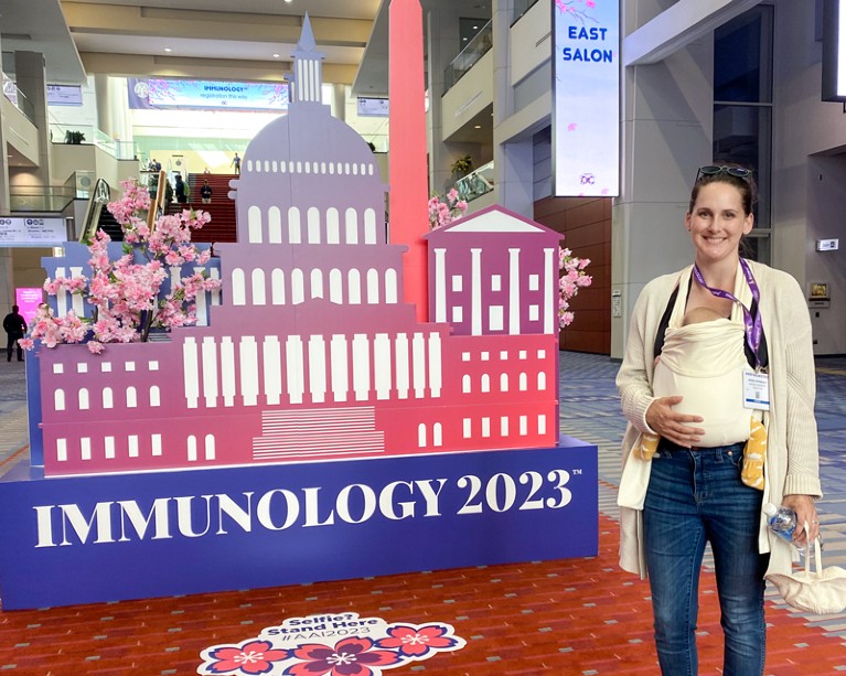 A woman with a baby in a front carrier stands beside a conference sign saying ‘Immunology 2023’